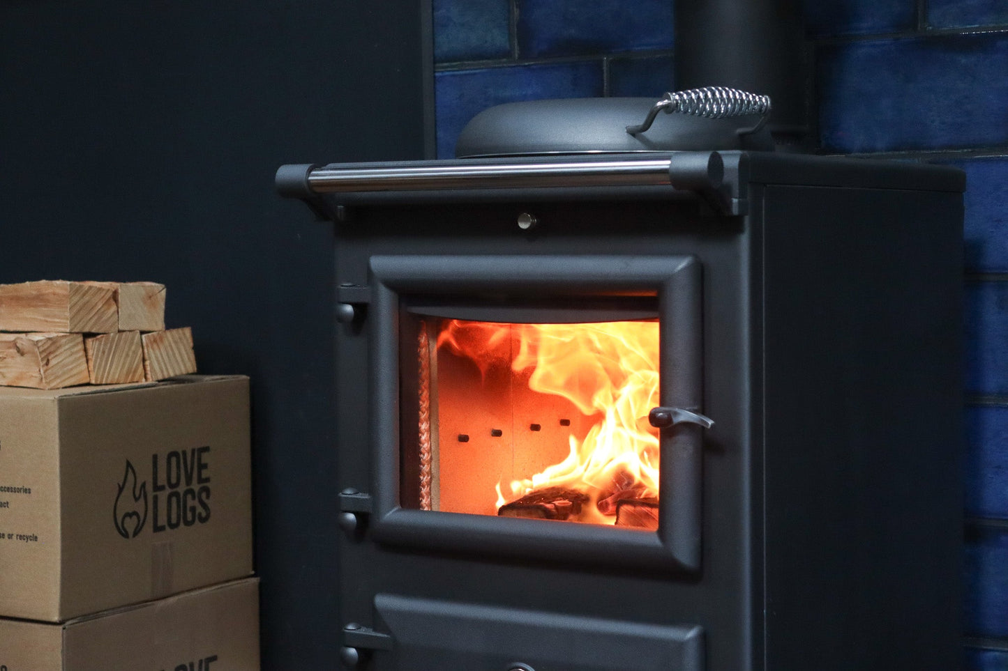 A blazing fire inside an Essie log burner with Love Logs boxes next to it and firewood on top of the boxes